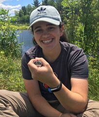 Olivia Rooney holds a nestling tree swallow. Olivia is wearing a gray hat and smiling.