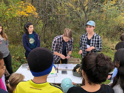 Three researchers stand in front of a table and demonstrate bird banding techniques while students watch.
