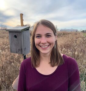 Dr. Jennifer Uehling stands in front of a nest box. She is wearing a purple shirt and is smiling.